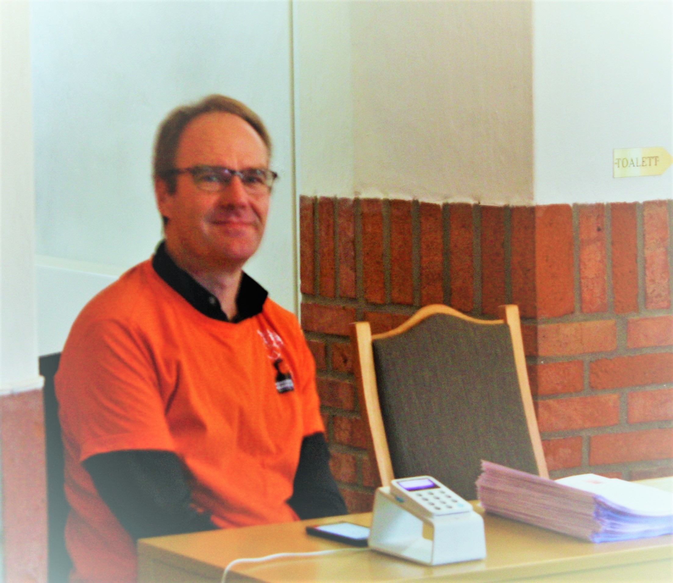 Mann med briller og oransje t-skjorte sitter med betalingsterminal og programhefter foran seg.